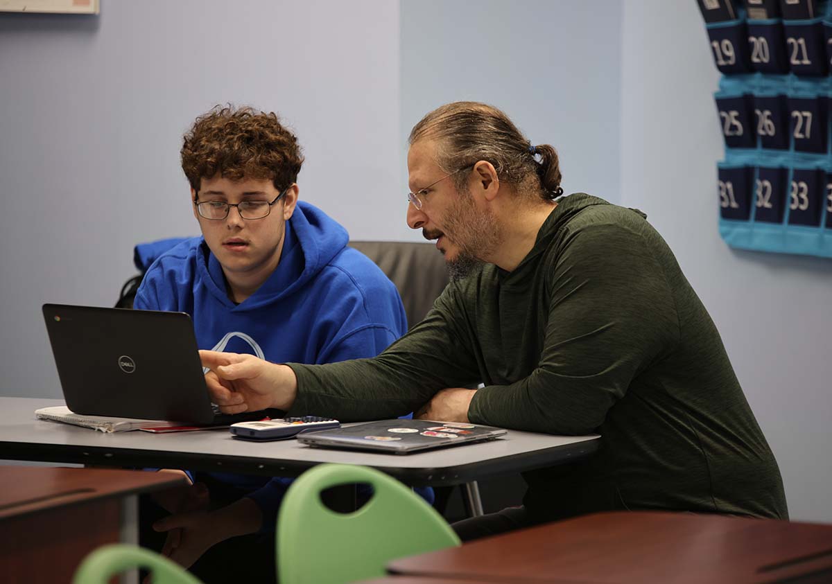 HSA Teacher smiles while kneeling beside a young student in a classroom setting.